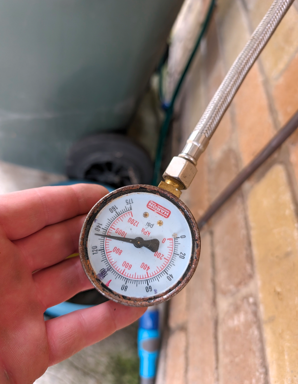 Hand holding a round pressure gauge attached to a braided hose near plumbing pipes, with a red needle visible — TasWide Plumbing in Kingston, TAS