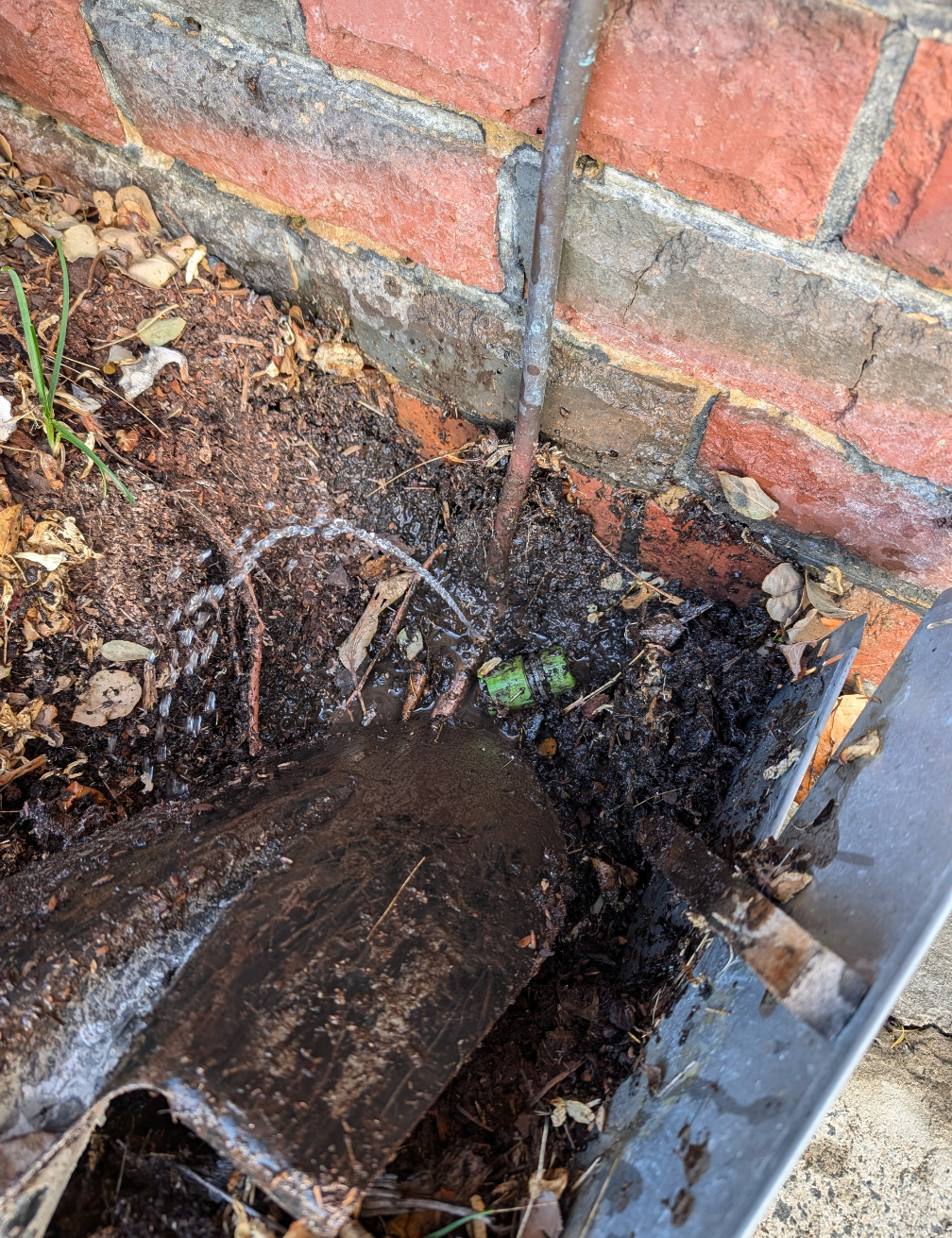Broken drain pipe and muddy debris at the base of a brick wall outdoors — TasWide Plumbing in Kingston, TAS