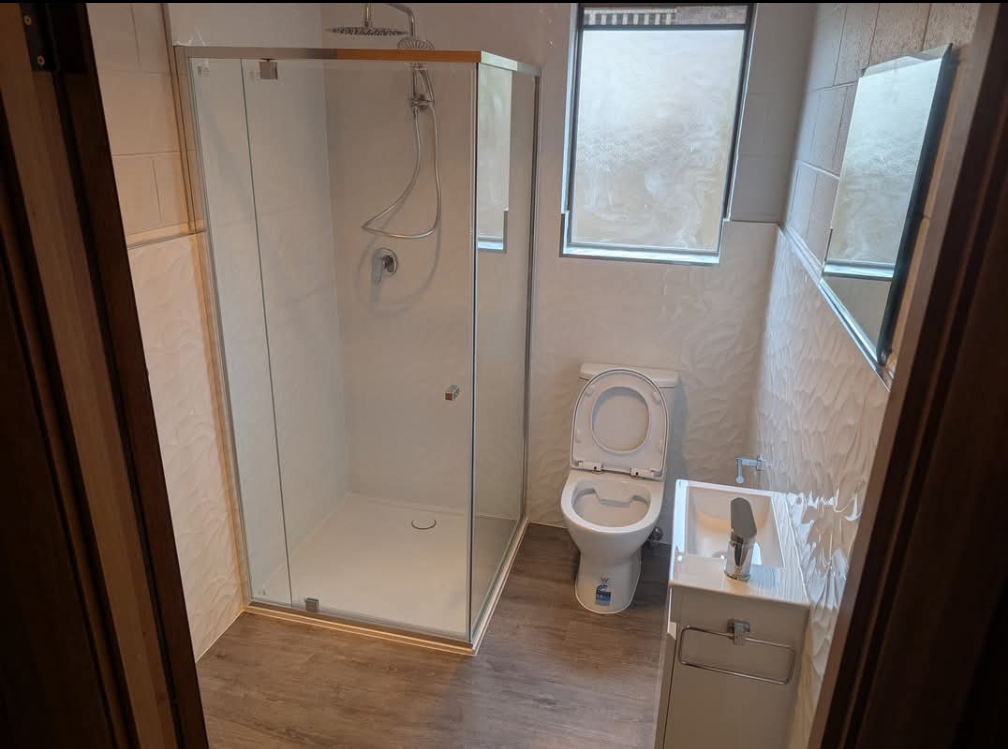 Small bathroom with shower, toilet, sink, and frosted window; toilet seat is open. — TasWide Plumbing in Kingston, TAS