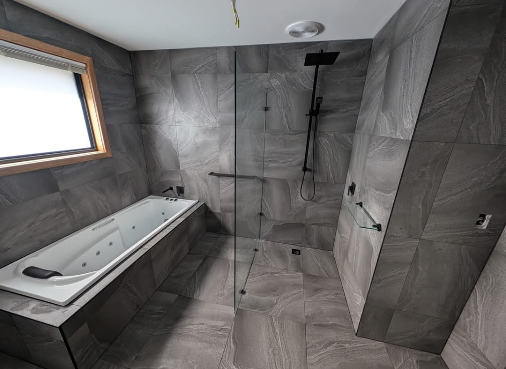 Modern gray-tiled bathroom with a white soaking tub, glass shower, and window. — TasWide Plumbing in Kingston, TAS