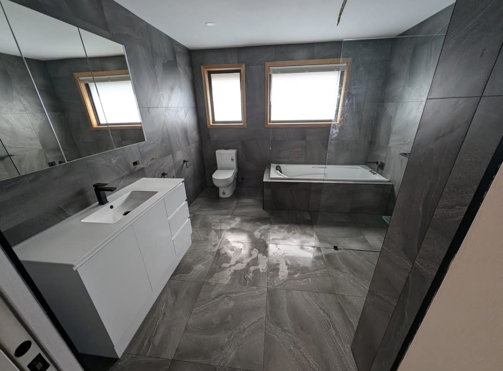 Modern bathroom with gray tile, white vanity, toilet, bathtub, and two windows — TasWide Plumbing in Kingston, TAS