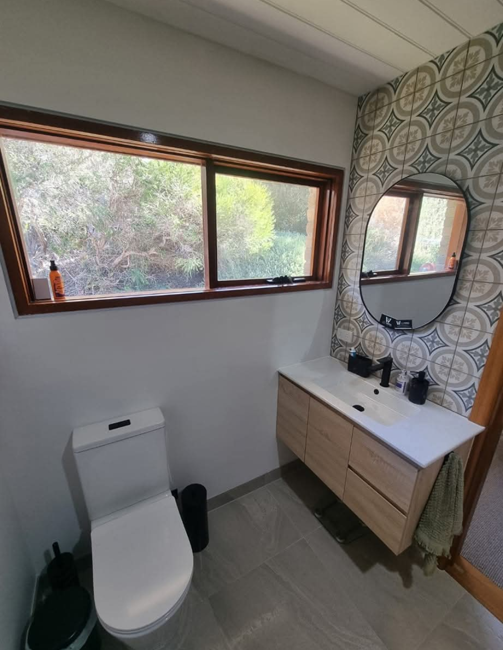 Small bathroom with toilet, vanity, mirror, and large window with patterned wallpaper. — TasWide Plumbing in Kingston, TAS