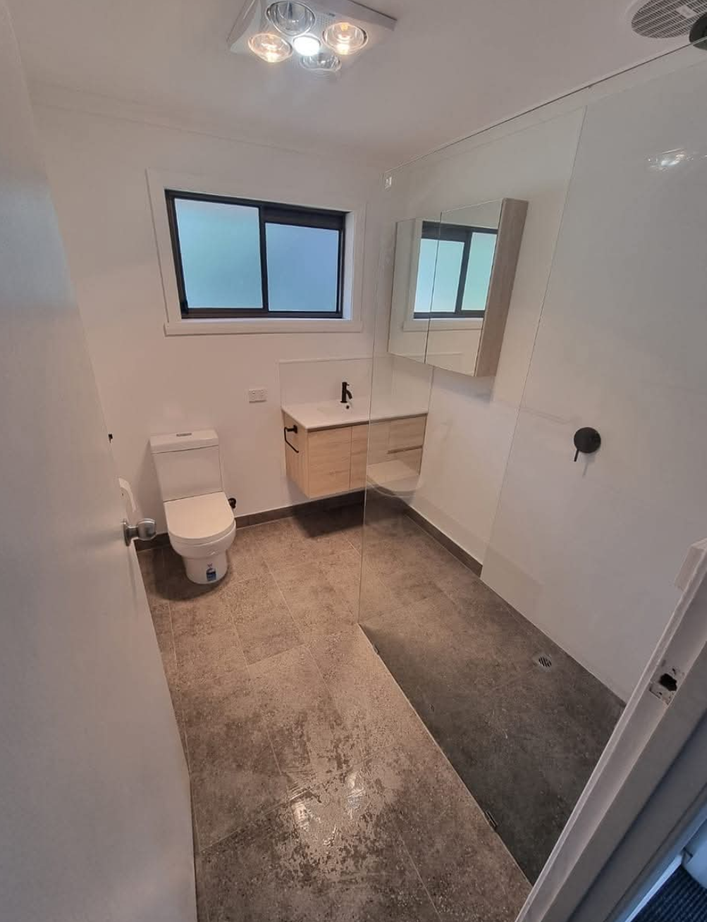 Small modern bathroom with toilet, floating vanity, large mirror, window, and tiled floor. — TasWide Plumbing in Kingston, TAS