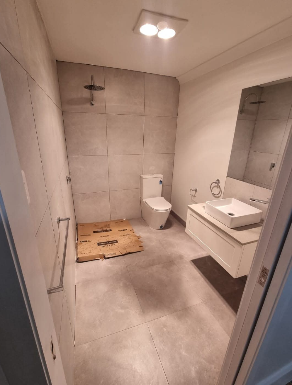 Small modern bathroom with gray tile, toilet, shower, sink, and mirror under ceiling lights. — TasWide Plumbing in Kingston, TAS