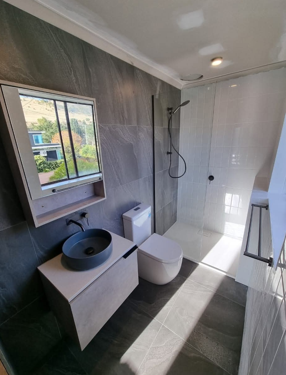 Modern bathroom with sink, toilet, shower, gray tile, and sunlight through a window — TasWide Plumbing in Kingston, TAS