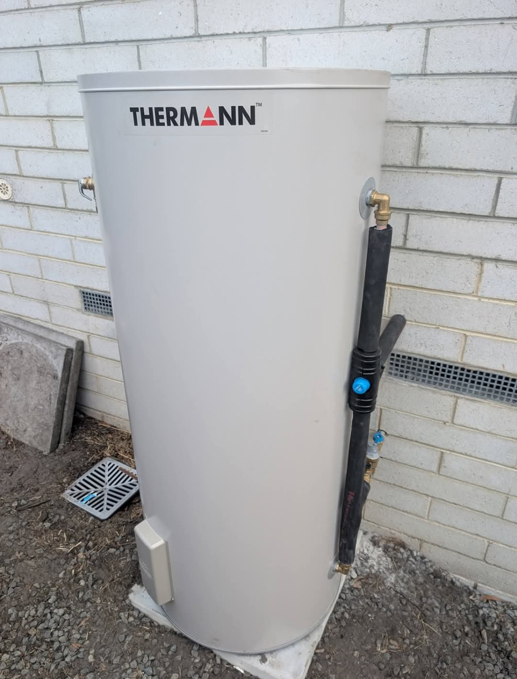 White Thermann hot water tank beside a brick wall, with exposed piping and controls outdoors. — TasWide Plumbing in Kingston, TAS