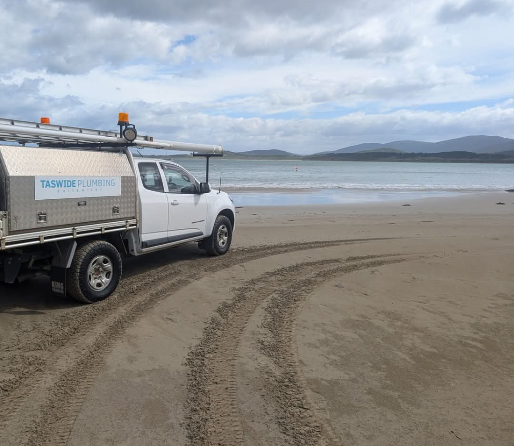 White utility truck parked on a beach with tire tracks in the sand and ocean under a cloudy sky. — TasWide Plumbing in Kingston, TAS