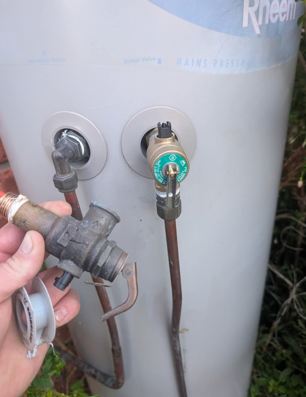 Water heater with two valves and a hand holding a pipe fitting beside it. — TasWide Plumbing in Kingston, TAS