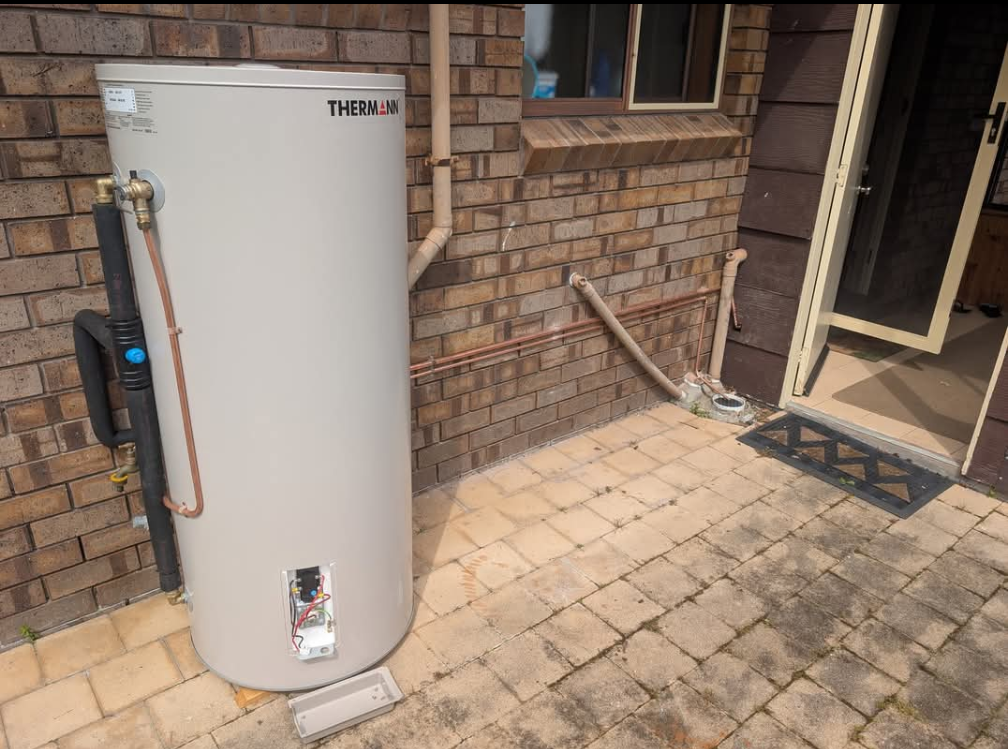 Outdoor gas water heater beside a brick house patio with open doorway and hose nearby — TasWide Plumbing in Kingston, TAS