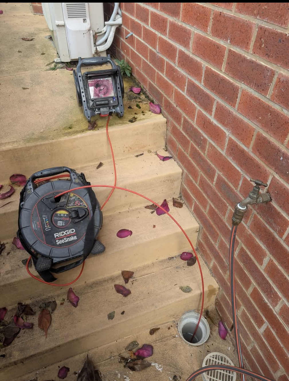 Red brick wall and concrete steps with a hose reel, extension cord, and fallen leaves outside a house — TasWide Plumbing in Kingston, TAS