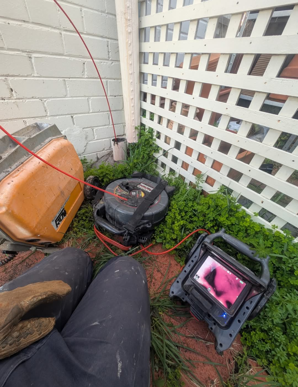 Outdoor corner with grass, lattice fence, orange container, black equipment, and red cord scattered on the ground — TasWide Plumbing in Kingston, TAS