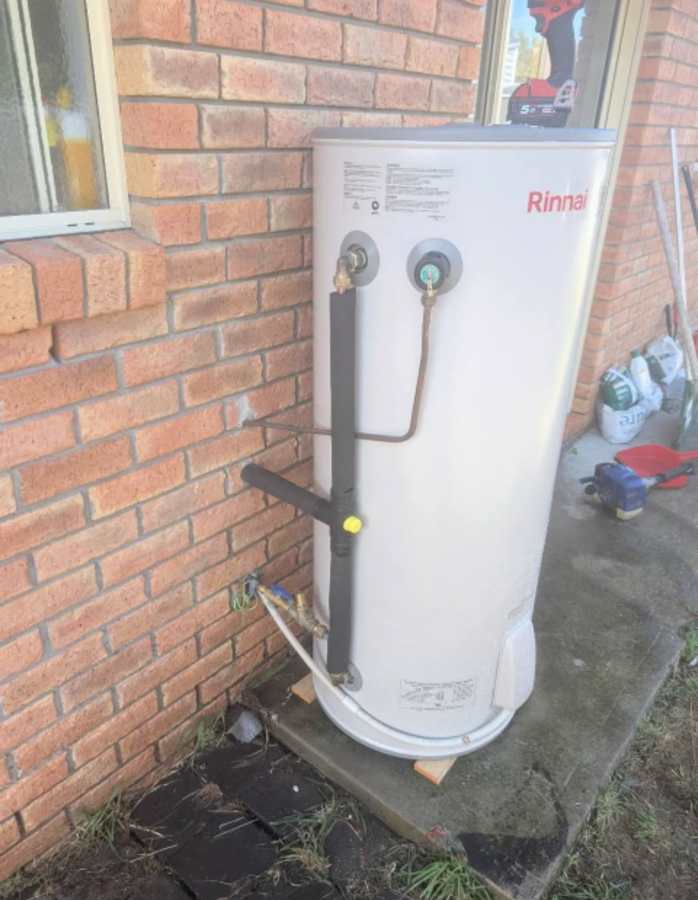 White Rinnai water heater beside a brick house, with pipes and valves attached outdoors. — TasWide Plumbing in Kingston, TAS