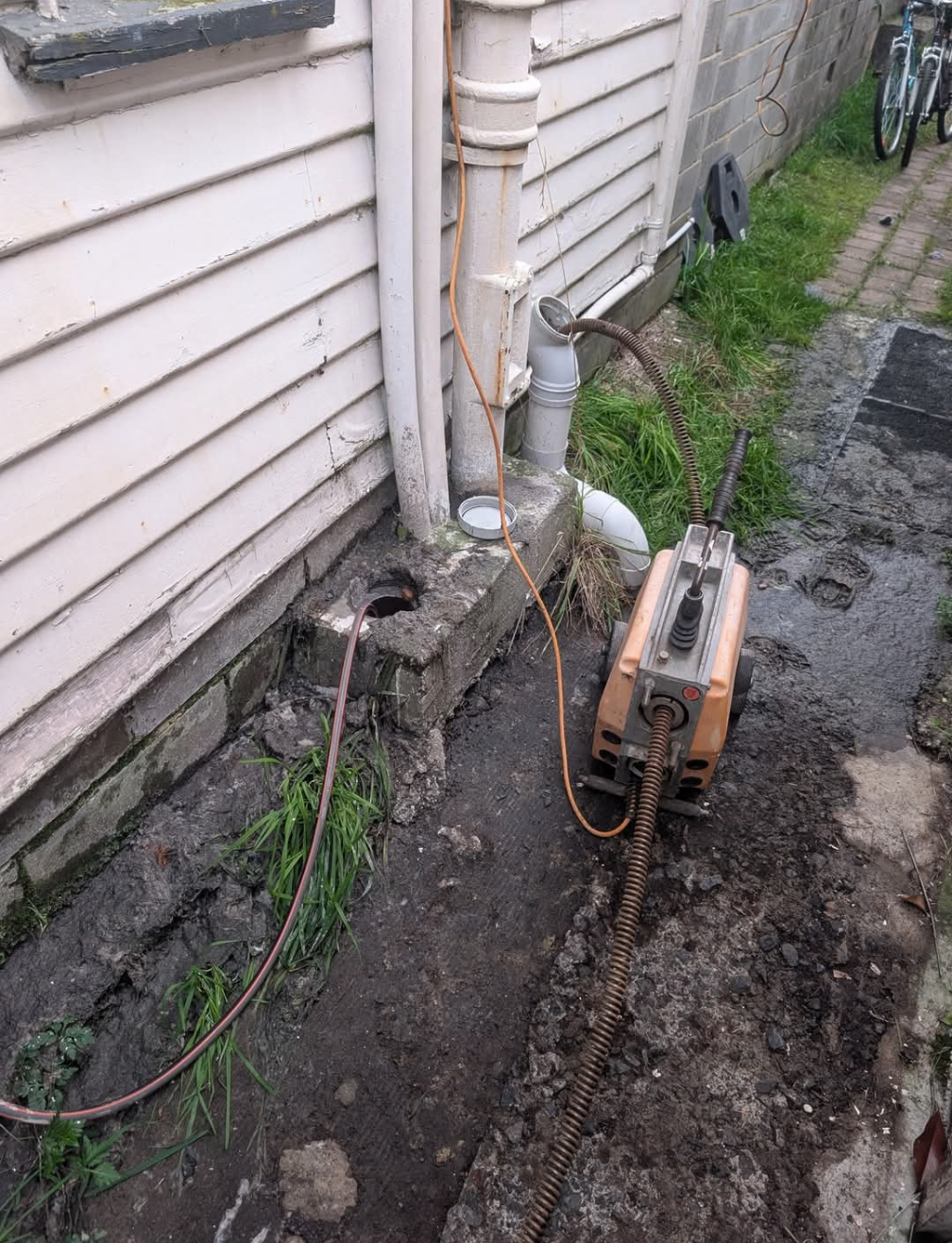 Outdoor side yard with a compressor, hoses, and wet ground beside a house wall and downspout — TasWide Plumbing in Kingston, TAS