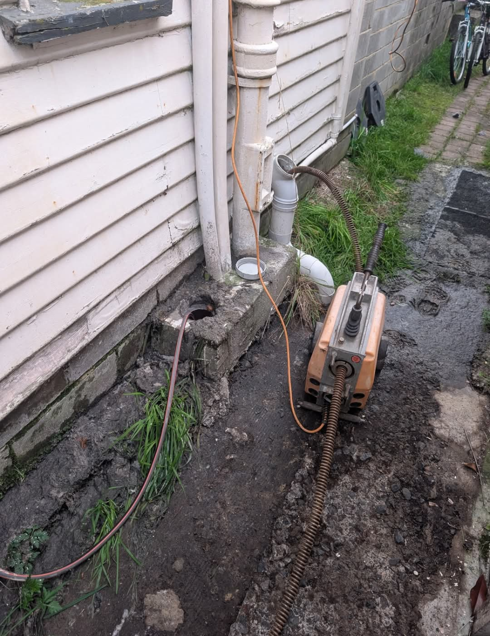 Orange tool on a muddy path beside a house wall and downspout, with cables and a hose nearby — TasWide Plumbing in Kingston, TAS
