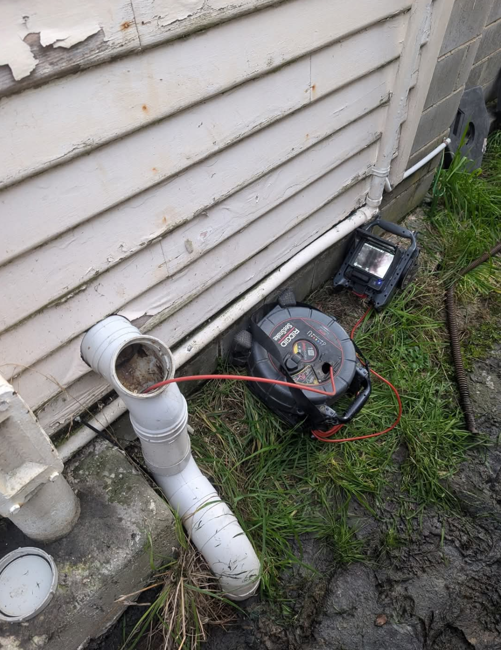 Broken drainage pipe beside house siding with a black blower and red extension cord on muddy ground — TasWide Plumbing in Kingston, TAS