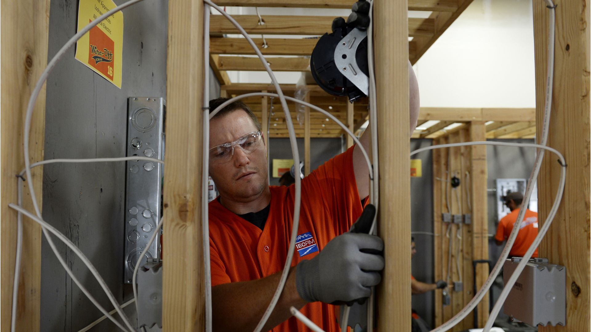 Worker in orange shirt installing wiring inside a wooden house frame.