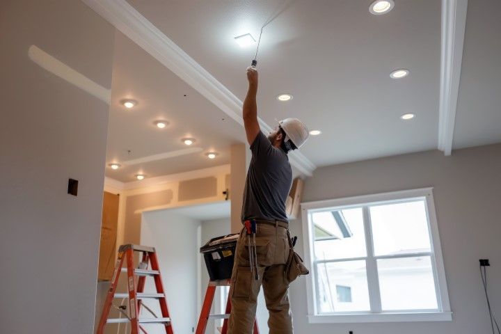 Worker installing a light fixture in a bright room with recessed ceiling lights and a ladder