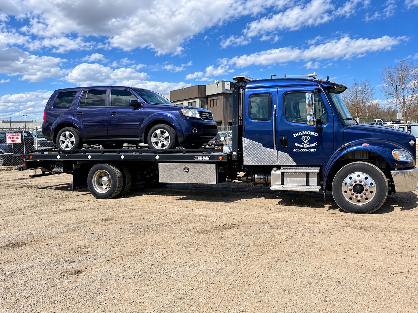 Blue tow truck carrying a dark blue SUV on a gravel lot under a partly cloudy sky