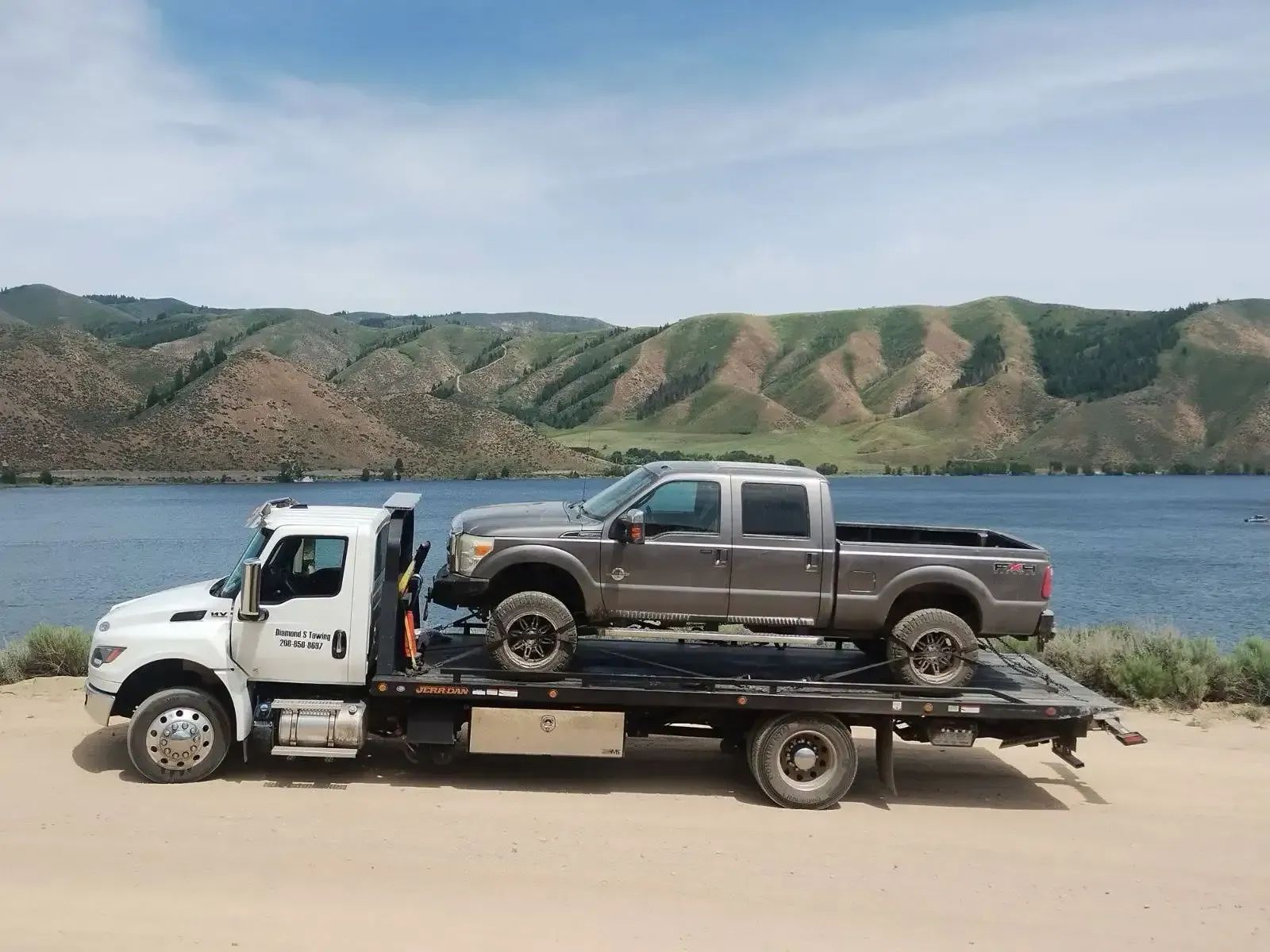 Tow truck hauling a blue pickup beside a lake with green hills in the background