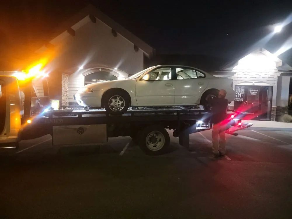 White car on a tow truck at night. A person stands beside the truck with lights on. Building in background.