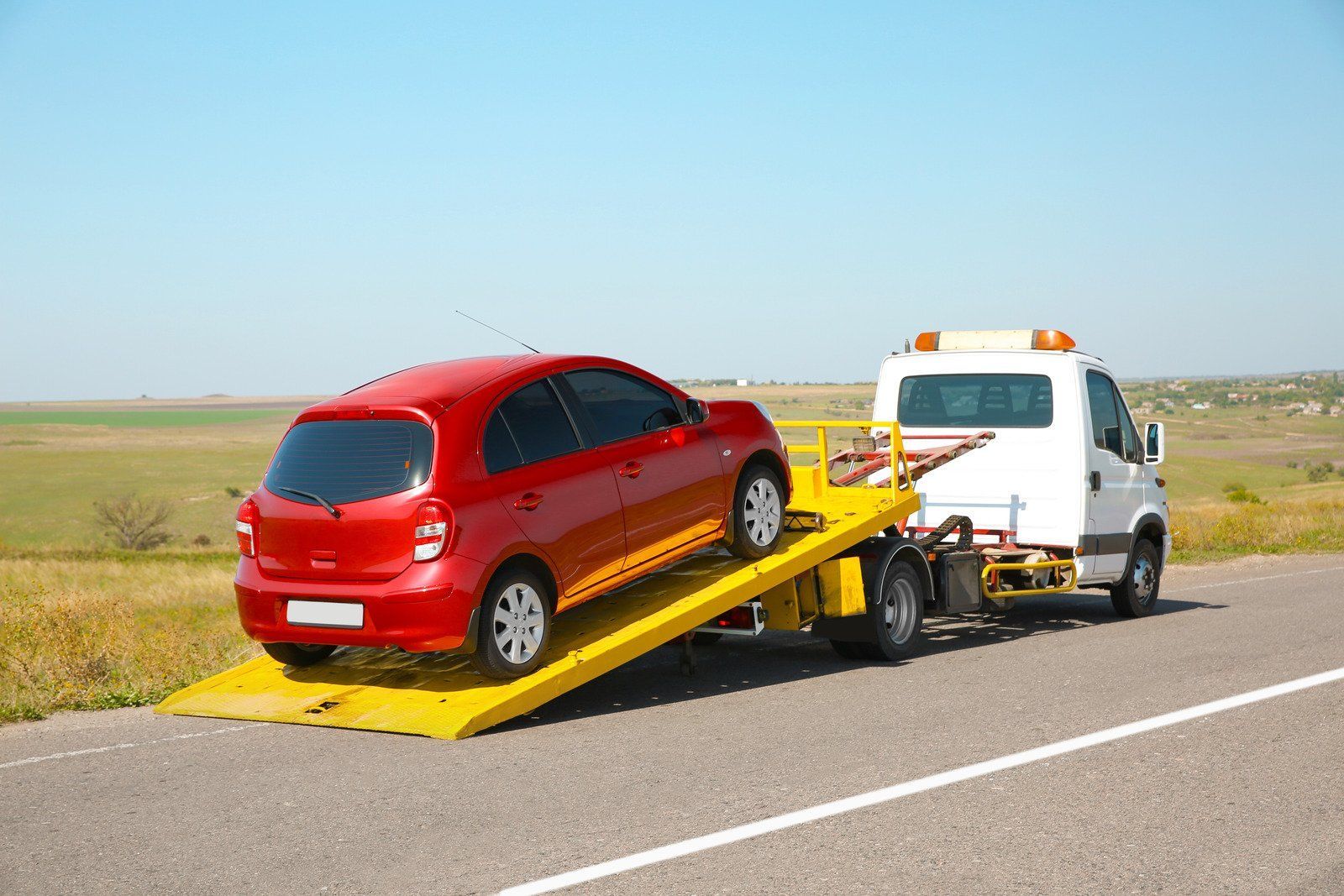 A red car being loaded onto a yellow flatbed tow truck on the side of a road against a clear blue sky.
