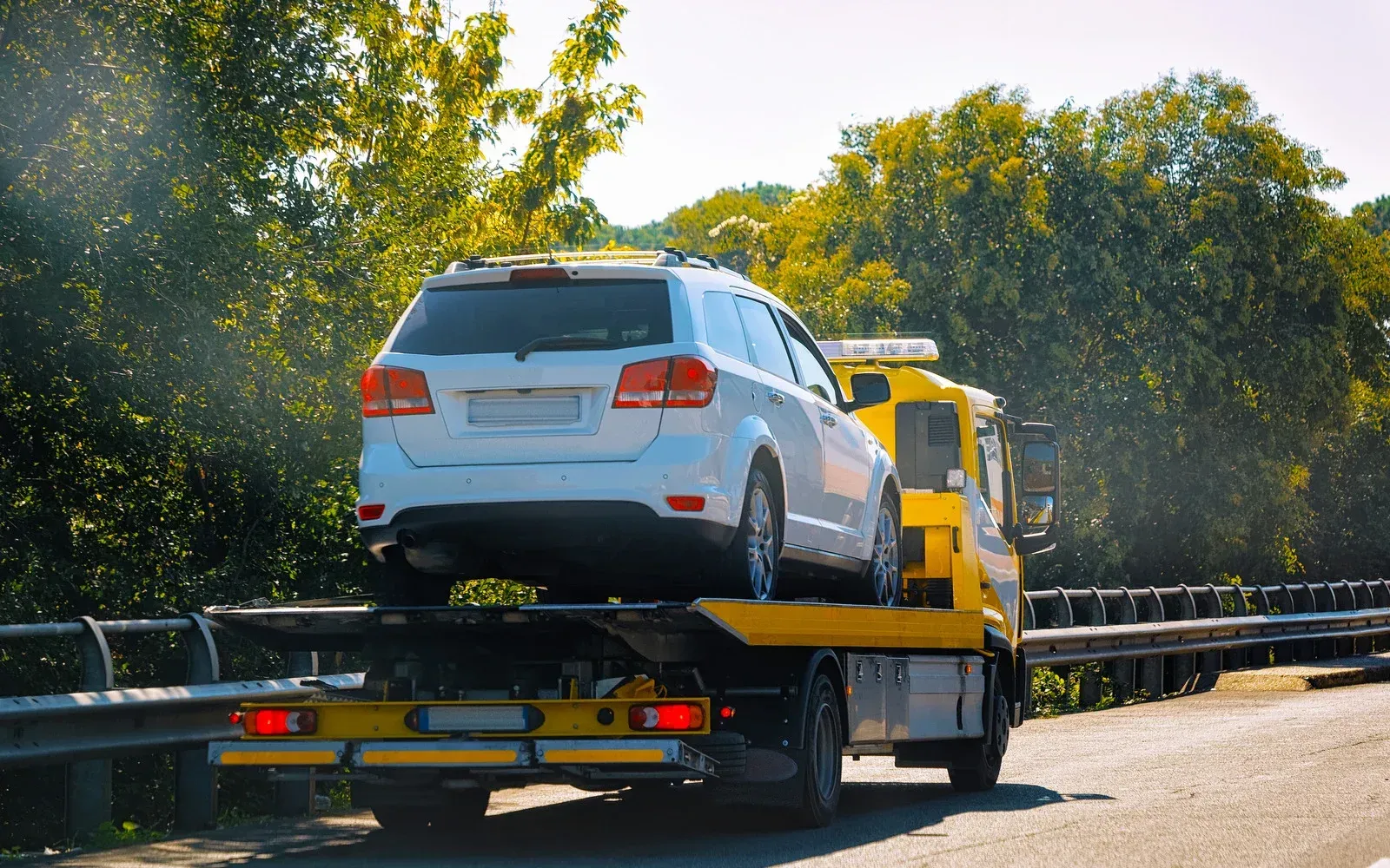 White SUV loaded on a yellow flatbed tow truck driving on a road with trees behind it