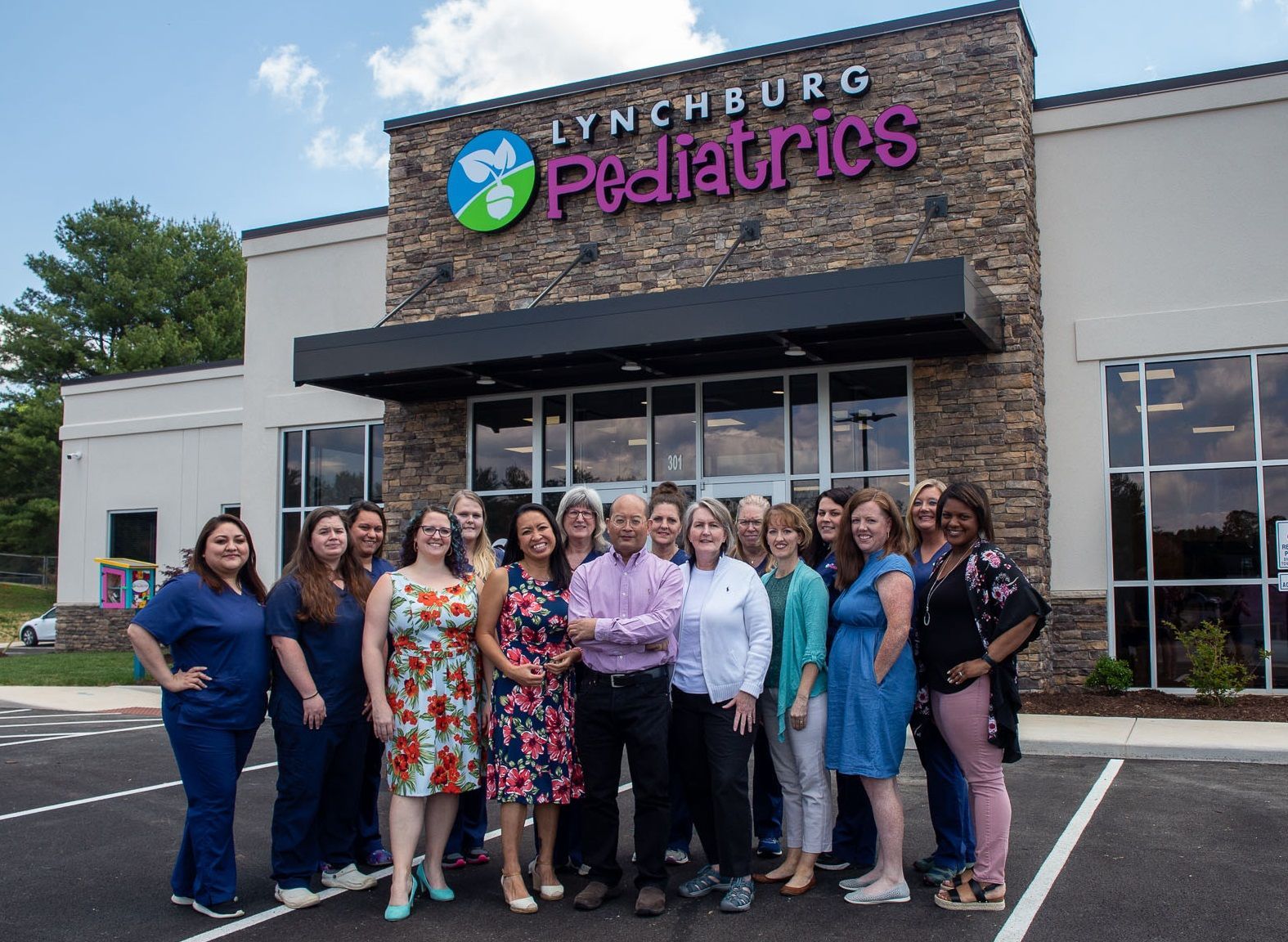 A large group of staff posing for a photo in front of the Lynchburg Pediatrics building on a sunny day.