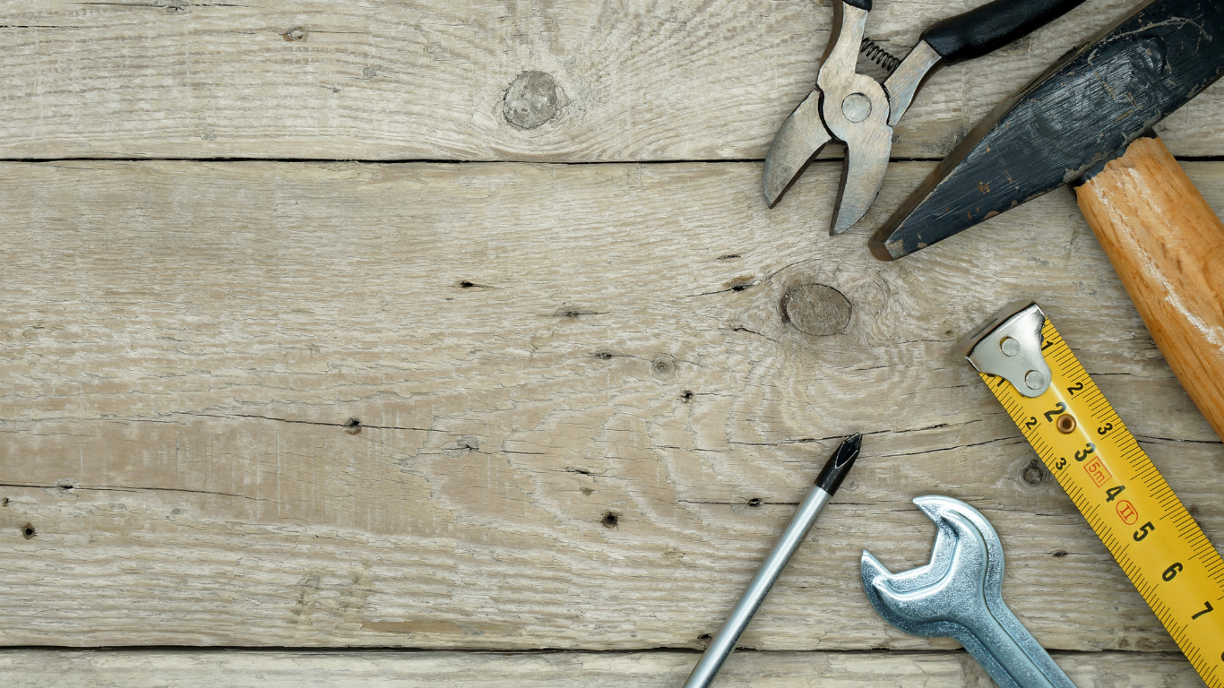 Tools on a rustic wooden workbench: pliers, wrench, screwdriver, hammer, and measuring tape.
