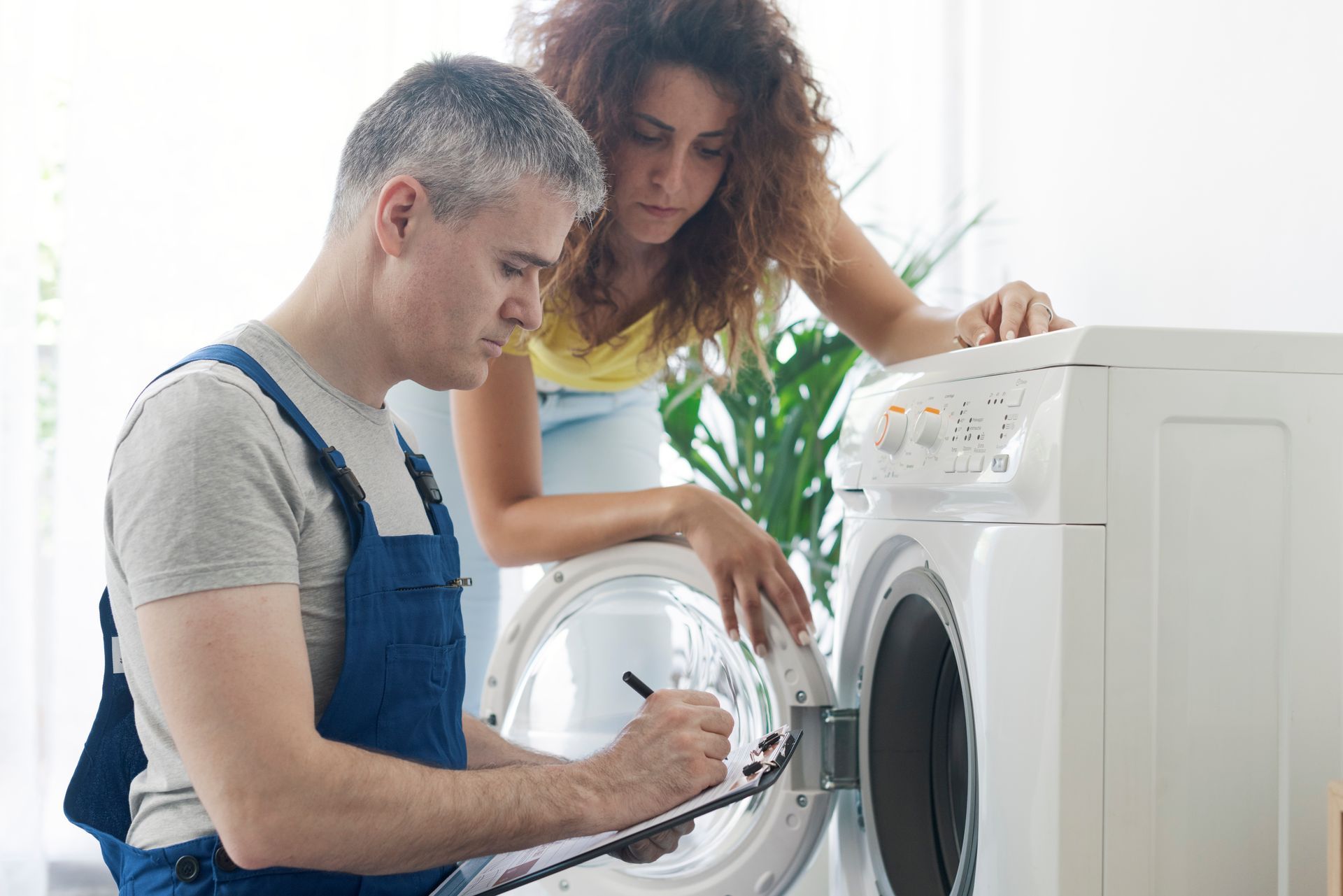 A technician is inspecting a washing machine while a homeowner looks on.