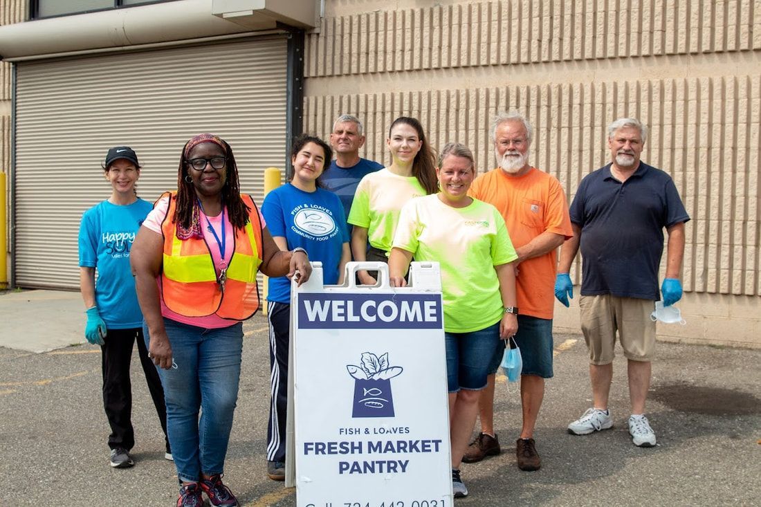 Group of volunteers standing behind a “WELCOME” sign outside a fresh market pantry