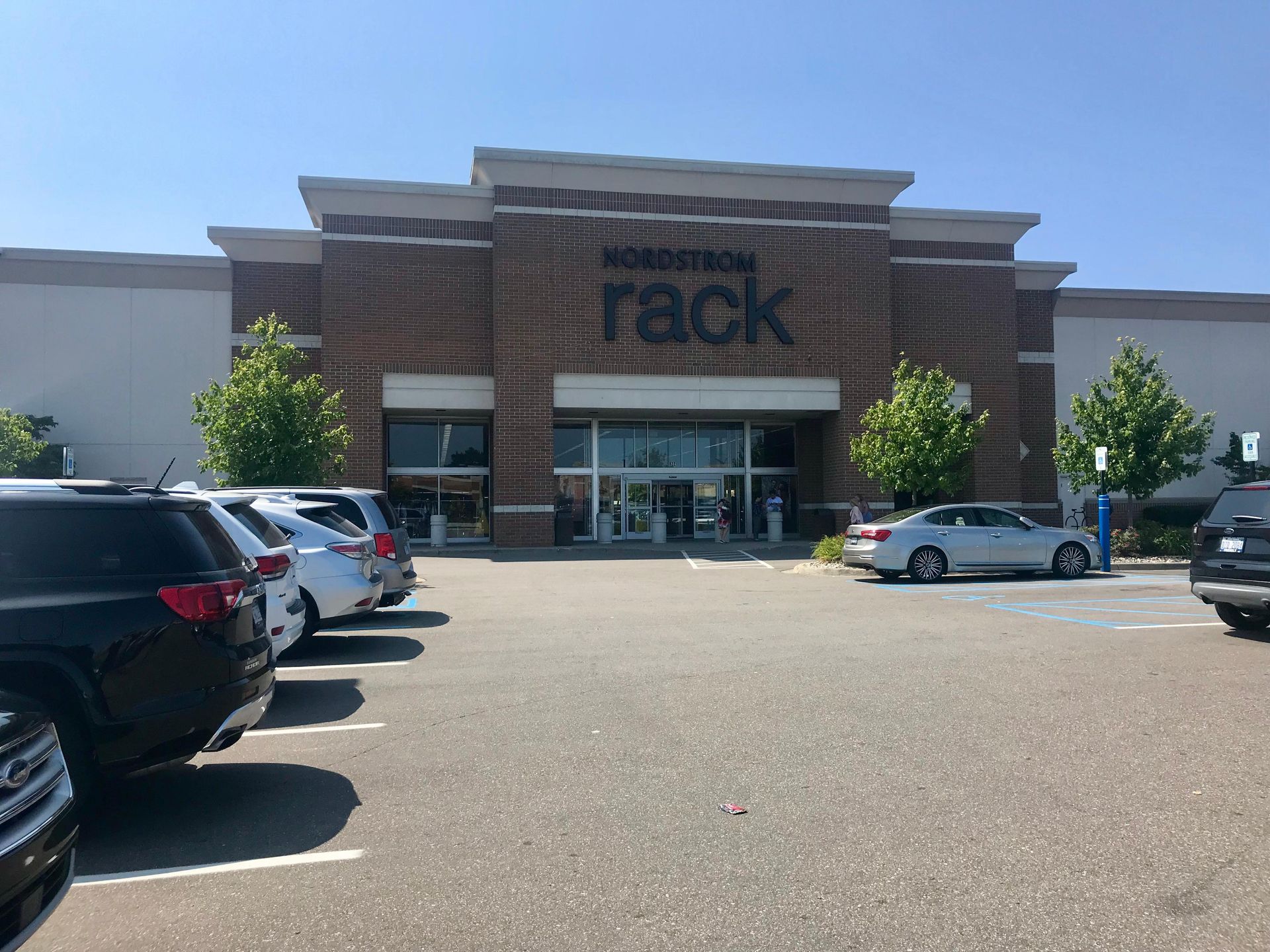 Front entrance of Nordstrom Rack store in a shopping center, with parked cars under a bright blue sky.