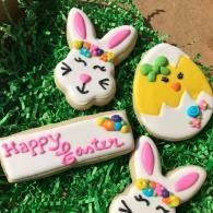 Easter-themed decorated cookies on grass, including a bunny face, egg, and “Happy Easter” cookie