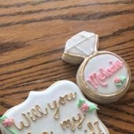 Decorated cookies on a wooden table, including a ring-shaped cookie and a “Will you be my bridesmaid?” cookie 