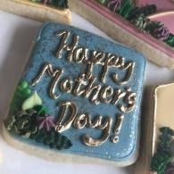 Blue square cookie with gold icing reading “Happy Mother’s Day”