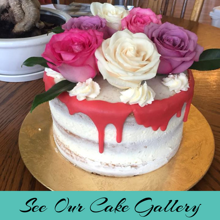 White drip cake topped with pink, cream, and purple roses on a gold board, with “See Our Cake Gallery” text below