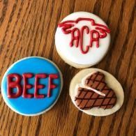 Three decorated cookies: blue “BEEF,” red “ACA,” and a brown steak on a wooden surface