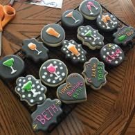 Decorated black iced base cookies with colorful cocktail and candy designs on them arranged on a wooden table