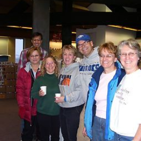 Group of people smiling, standing indoors near boxes. Some hold cups, wearing casual clothes.