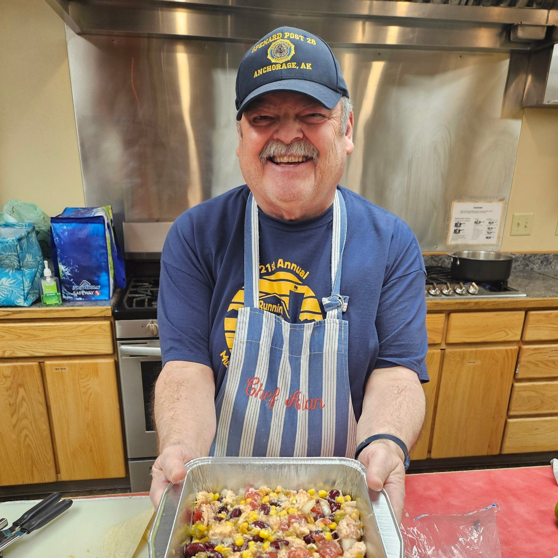 Smiling person holding pan of food in a kitchen. Wearing hat, apron. Stainless steel appliances in background.