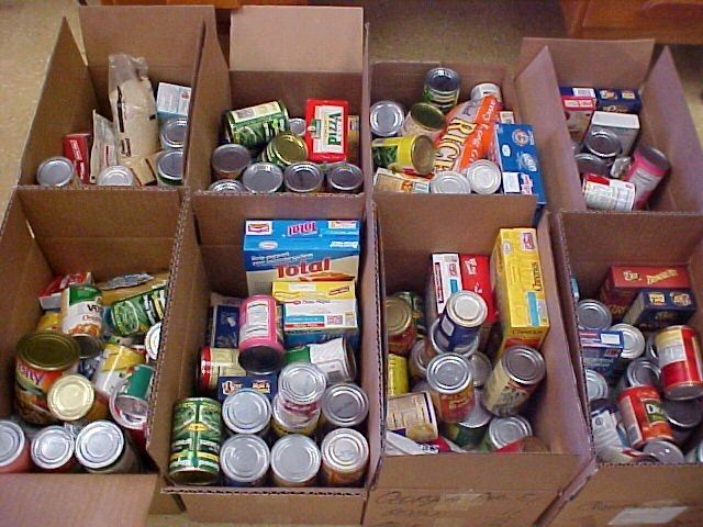 Boxes of food stacked on a table, likely a donation for a food drive, with additional food items on the floor.