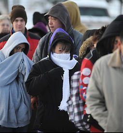 People bundled in winter clothing outdoors, some with scarves covering faces, in a cold weather setting.