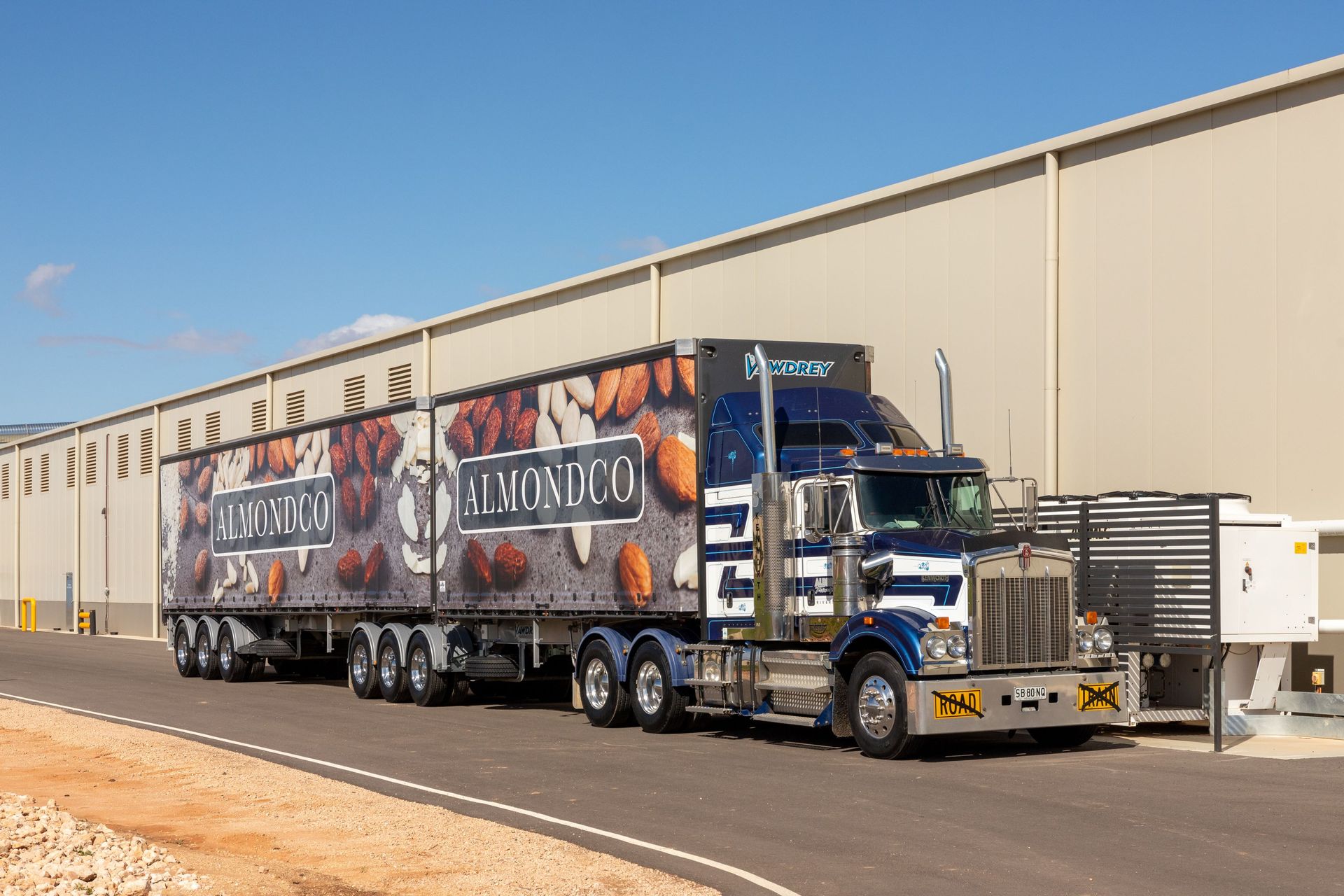 Blue semi-truck with a graphic trailer parked beside a beige warehouse on a sunny day
