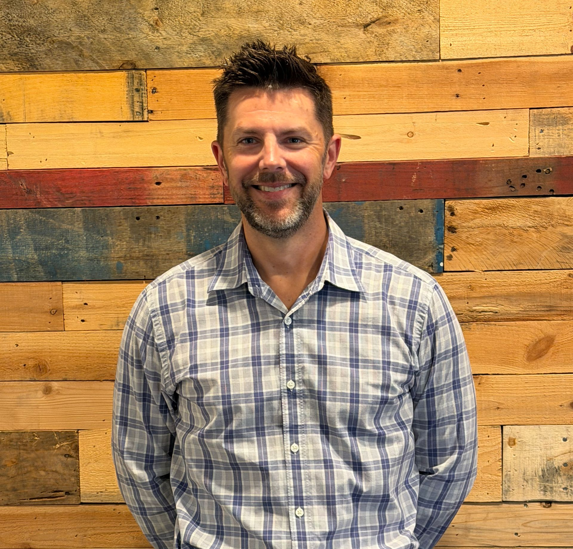 Man in blue shirt with a small beard smiles in front of a wood-paneled wall.