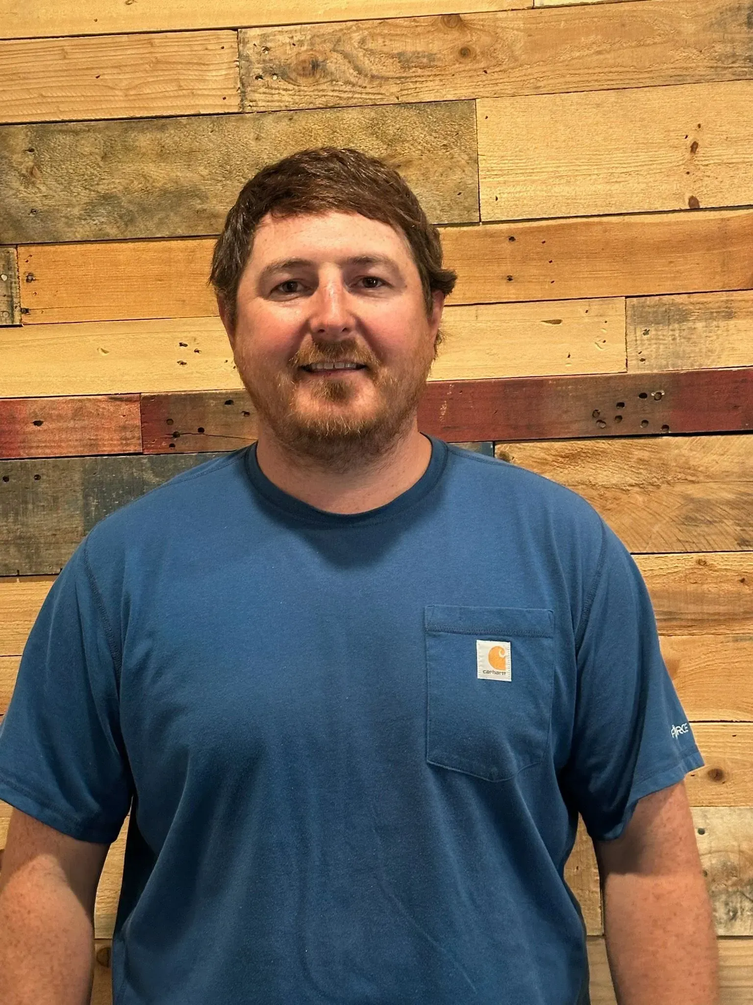 Man in blue shirt with a small beard smiles in front of a wood-paneled wall.