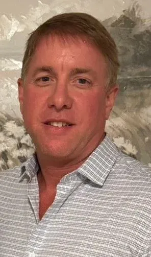 Person in a black blazer and brown button-up shirt smiling. They have curly hair and are indoors against a beige wall.