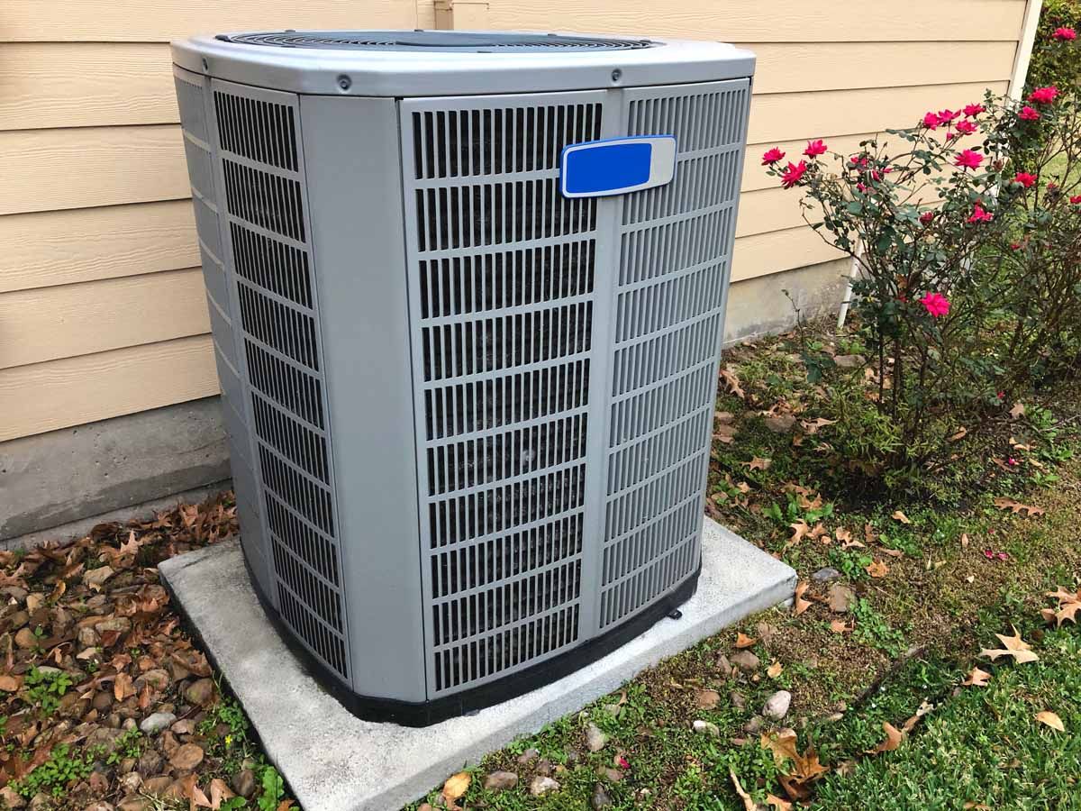 A gray, outdoor residential air conditioning unit sitting on a concrete pad next to a house with rose bushes.
