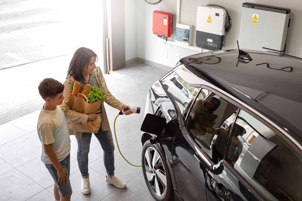 A person carrying groceries plugs a charging cable into an electric car inside a garage with wall-mounted power units.