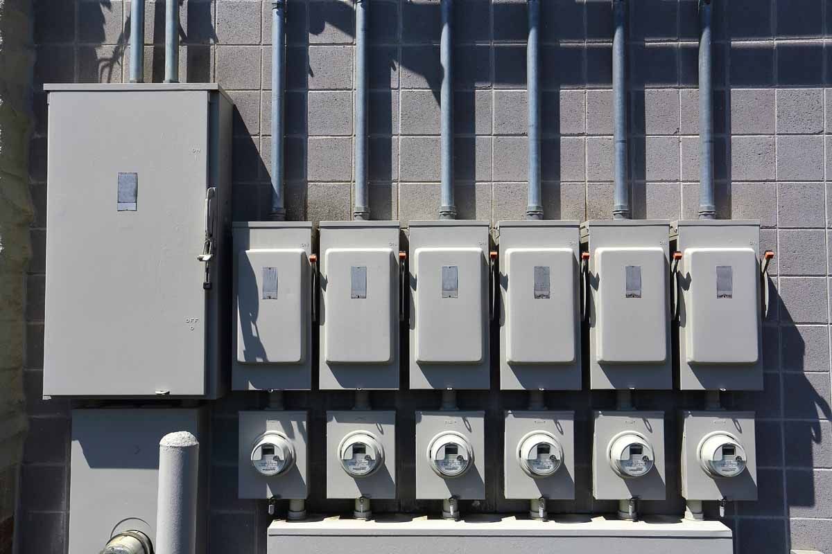 A bank of six electrical meters and associated disconnect boxes mounted on a gray concrete block wall.