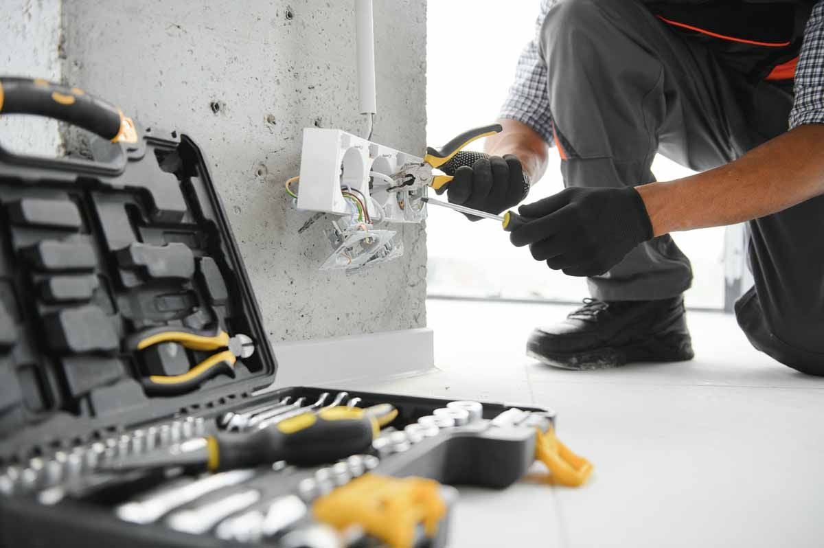 An electrician wearing black gloves uses a screwdriver to work on a white wall-mounted electrical outlet near a tool kit.