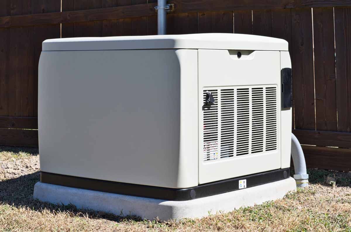 A beige standby home generator sits on a concrete pad in front of a wooden fence on a patch of dry grass.